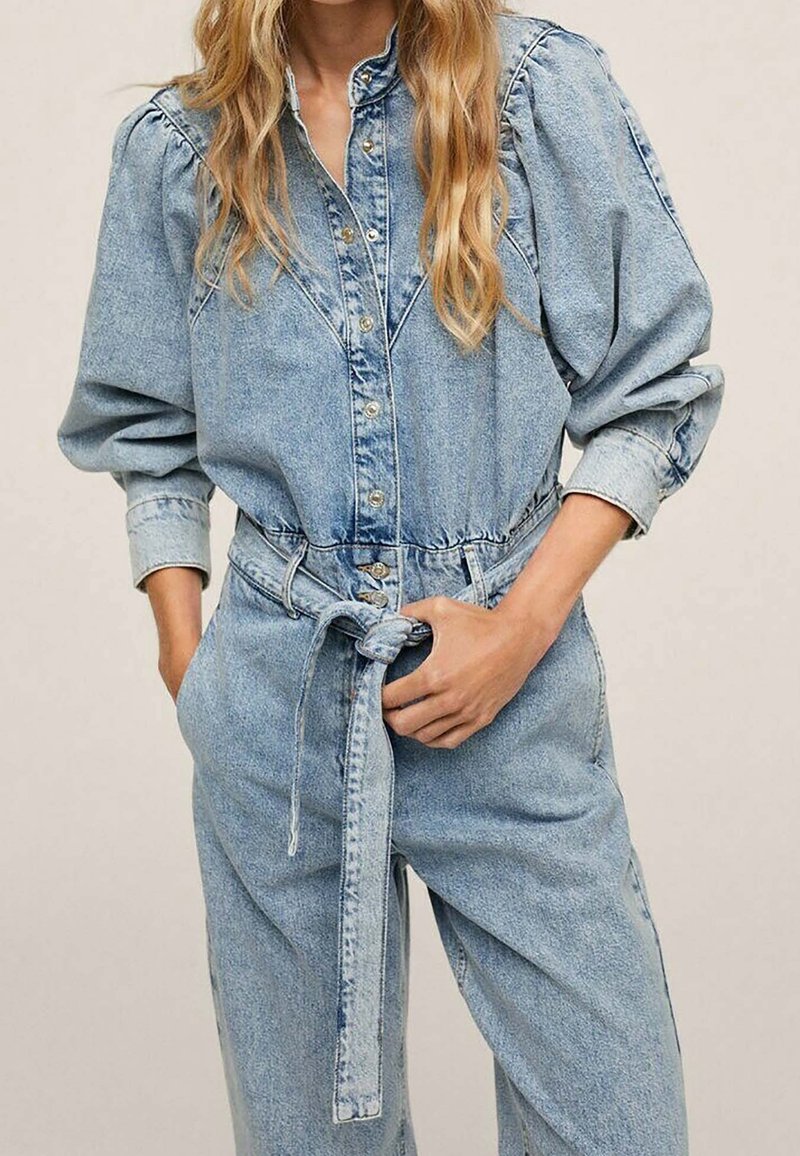 Femme portant une combinaison en denim bleu clair avec des épaules bouffantes, une fermeture à boutons sur le devant et une ceinture nouée à la taille, debout les mains près des poches.
