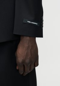 Κοντινό πλάνο ενός σκουρόχρωμου χεριού που φοράει μια μαύρη σακάκι με ετικέτα Karl Lagerfeld ευδιάκριτη στο μανίκι.