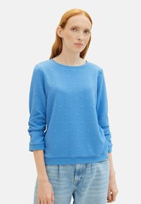 Felpa blu con texture e collo a giro, caratterizzata da piccoli motivi a cuore in rilievo e maniche lunghe, abbinata a jeans azzurri chiari.