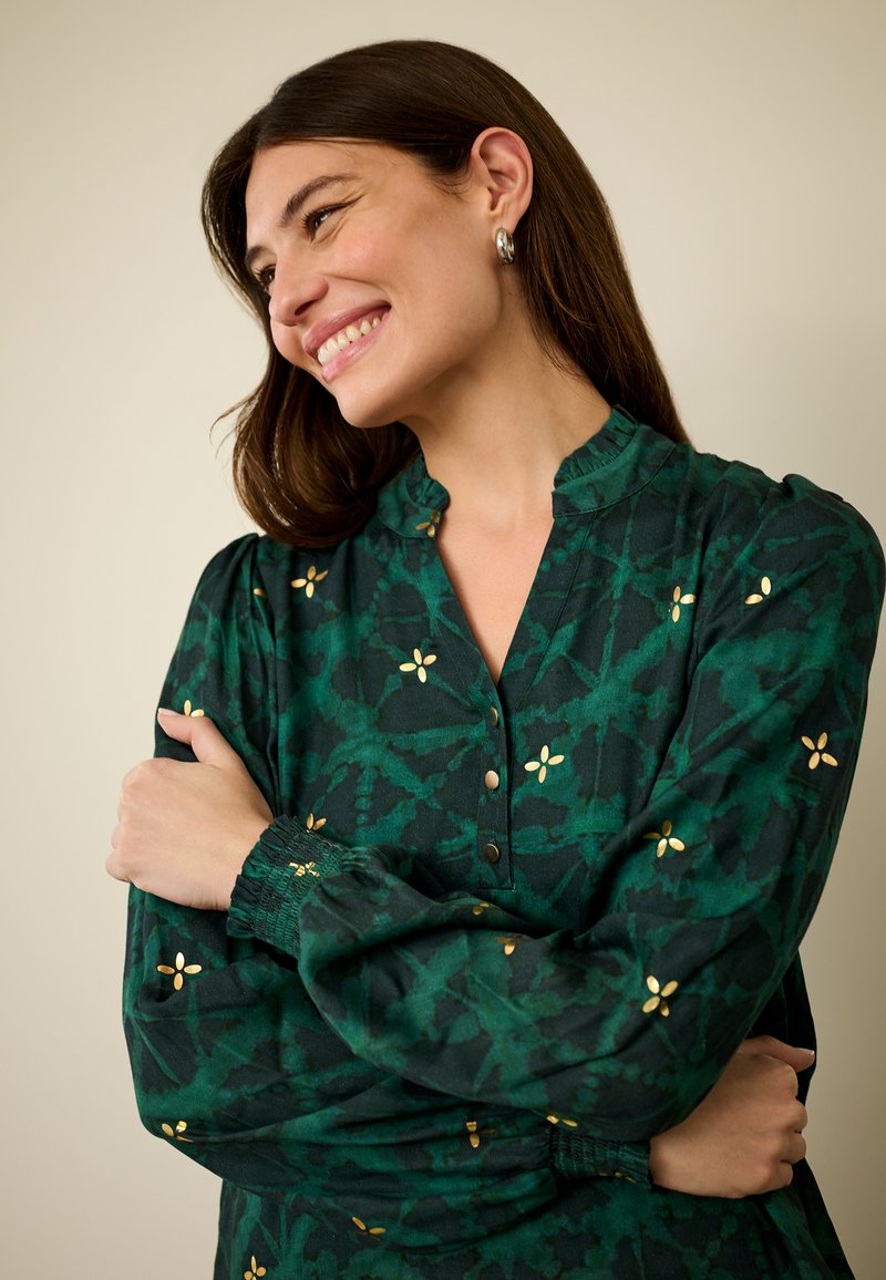 Žalia raštuota palaidinė su raukiniais apykaklėje, papuošta auksinėmis gėlių detalėmis ir sagomis priekyje. Tekstūruota medžiaga su suraitytais rankogaliais.