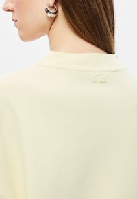 Šviesiai geltonas mezgimo megztinis su dygsniuotu apykakliu. Ant nugaros yra subtiliai išsiuvinėtas logotipas, o audinys yra lygios tekstūros.