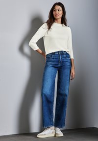Weißes geripptes Langarmoberteil kombiniert mit hoch taillierten, blauen weiten Jeans. Das Model trägt weiße Sneakers und steht vor einem grauen Hintergrund.
