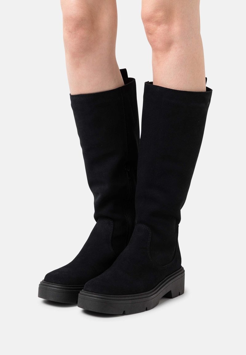 Jambes portant des bottes mi-mollet en daim noir avec des semelles épaisses en caoutchouc et des talons bas à blocs épais sur un fond clair uni.
