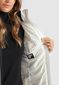 Veste blanche avec une texture lisse et une fermeture éclair, tenue ouverte pour révéler un intérieur beige. Présente une étiquette en tissu noir sur la doublure gauche.