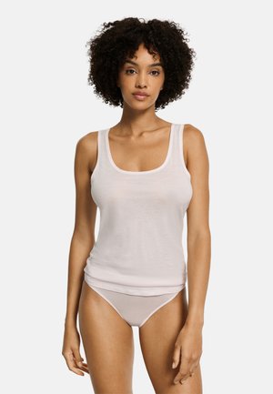 Vrouw met krullend haar draagt een wit hemdje en bijpassend ondergoed, staand met het gezicht naar voren tegen een effen lichte achtergrond.