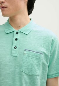 Miętowa koszulka polo z teksturowanej tkaniny, z trzema czarnymi guzikami i kieszenią naszywaną z kontrastowymi paskami.