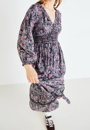 Femme portant une longue robe noire à fleurs roses et blanches, soulevant légèrement la jupe, assortie à des baskets noires et blanches.