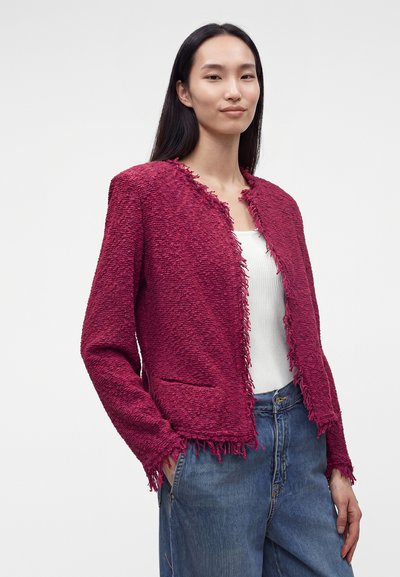 Femme aux longs cheveux foncés portant une veste bordeaux texturée sur un haut blanc et un jean bleu, debout devant un fond clair et uni.
