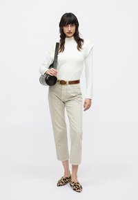 Λευκό μακρυμάνικο τοπ, μπεζ παντελόνι ψηλόμεσο, καφέ ζώνη, μαύρη τσάντα με υφή και παπούτσια με animal print. Το μοντέλο στέκεται μπροστά σε λευκό φόντο.