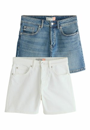Kvinde iført hvide denimshorts og en kort orangestribet top, med hånden hvilende i shorts-lommen udendørs nær et hegn.