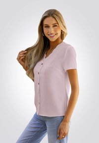 Camisa de manga corta, de color rosa pálido, con botones y una superficie texturizada, con un escote con forma de ondas, combinada con jeans de color azul claro.
