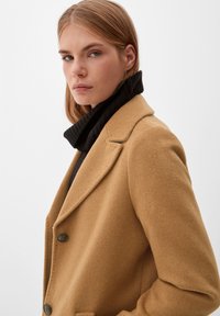 Manteau en mélange de laine camel avec un col cranté, deux boutons et des poches latérales, superposé à un pull à col roulé noir en maille côtelée.