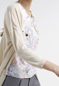 Ljusbeige kofta över en blommig topp i pastellfärger. Toppens halsringning är rund och tyget flödar. Gråbyxor syns.