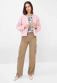 Lichtroze bomberjack met strakke manchetten, gedragen over een patroonblouse. Tan cargobroek met zijzakken. Beige puntige schoenen.