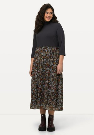 Schwarzes Ripp-Top mit hohem Kragen und 3/4-Ärmeln, kombiniert mit einem langen Rock mit Blumenmuster in verschiedenen Farben. Abgerundet mit Knöchelstiefeln.