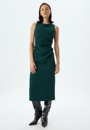 Vestido sin mangas verde con una silueta ajustada y detalle drapeado; combinado con botas negras hasta la rodilla con punta afilada y acabado texturizado.