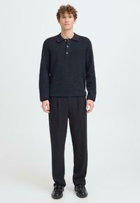 Junger Mann steht da und trägt einen dunklen, geknöpften Kragenpullover, lockere schwarze Hosen und schwarze Slipper vor einem schlichten, hellen Hintergrund.
