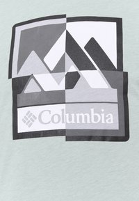 Camiseta verde claro con un diseño gráfico de formas geométricas en blanco y negro y el logo de "Columbia" en la parte inferior.
