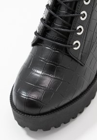 Bota de piel sintética de cocodrilo negra con punta redondeada, suela gruesa y estriada, y ojales de metal con cordones para el ajuste.