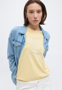 Νεαρή γυναίκα με κίτρινη ριγέ μπλούζα "Atelier 2010 Mos Mosh", ανοιχτόχρωμο τζιν μπουφάν και άσπρο παντελόνι, σε απλό φόντο.