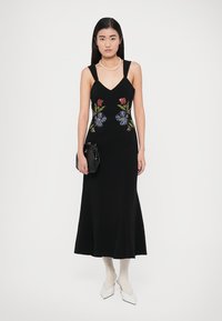 Rochie neagră fără mâneci, cu corset strâmt, broderie florală roșie și albastră pe partea frontală și tiv evazat. Model care ține o geantă mică neagră.