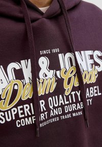 Bordeaux hættetrøje med snoretræk. Har stor hvid og guld tekst, der angiver "Jack & Jones Denim Goods" og "Superior Quality Label."