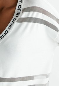 Top bianco con scollo a V, caratterizzato da strisce orizzontali grigie trasparenti e un colletto decorato con logo. Texture liscia, design aderente.