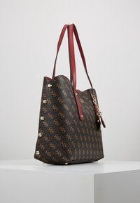 Bruine totebag met een logo patroon, rode leren hengsels en metalen accenten. Het ontwerp is ruim met zijstuds voor stabiliteit.