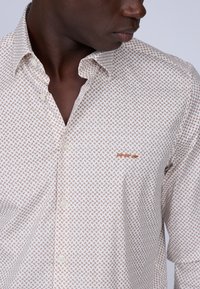 Chemise blanche avec un petit motif géométrique marron, boutonnière devant et un logo subtil brodé sur la poitrine. Tissu en coton, col classique.