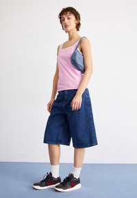Rosa gerippter Tank-Top mit weißen Akzenten, kombiniert mit lockeren blauen Jeansshorts und schwarzen Sneakers mit grünen Highlights. Weiße Socken sichtbar.