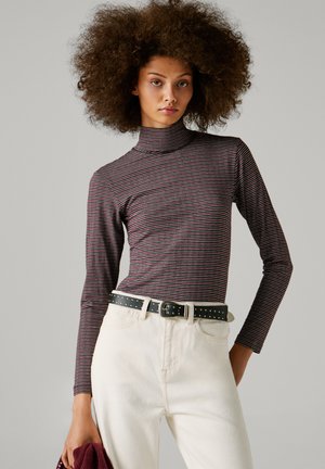 Vrouw met volumineus krullend haar, gekleed in een gestreepte coltrui en witte broek met een zwarte gesp met studs, holding een opgevouwen bordeauxkleurig kledingstuk.