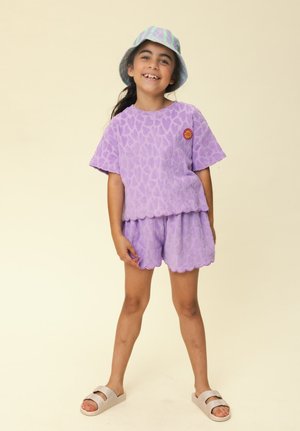 Smiling child wearing a purple textured shirt and shorts set, striped bucket hat, and beige slide sandals, standing against a plain background.