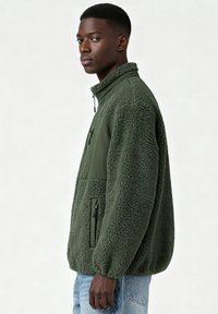 Veste polaire vert olive avec une texture douce, col à fermeture éclair partielle et poches latérales. Comprend un panneau en tissu contrastant sur la poitrine.