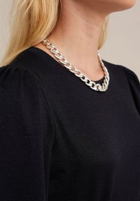 Sudraba ķēdes kaklarota uz tumša auduma blūzes ar savilktiem piedurknēm; kaklarota ir veidota no lielām, savstarpēji savienotām saitēm ar pulētu virsmu.