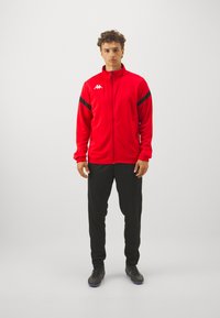 Veste de sport rouge avec des accents noirs, fermeture �éclair intégrale et col montant, associée à un pantalon noir et des chaussures de sport noires. Texture de tissu lisse.