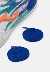 Zapatilla deportiva colorida con parte superior de piel blanca y acentos en teal, naranja y azul, acompañada de dos cordones azules.