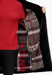 Černý vnější kabát s vzorovaným, kostkovaným interiérem. Interiér obsahuje designy houndstooth a plaid v červené, béžové a černé barvě. Vidět červený svetr.