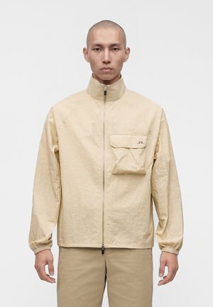 Homme portant une veste beige zippée avec un col montant et une grande poche poitrine, debout devant un fond blanc uni.