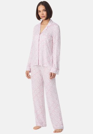 Femme debout pieds nus portant un ensemble de pyjama à manches longues à boutons avec un motif rose et blanc, comprenant un haut et un pantalon, sur un fond uni.
