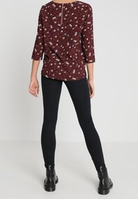 Blusa floreale bordeaux con maniche a tre quarti e dettagli con zip sul retro, abbinata a jeans neri aderenti e stivaletti alla caviglia.