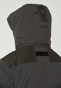 Mörkgrå jacka med en stor huva, med en matt yta och en kontrasterande svart axelsection. Inkluderar en etikett på huvan.