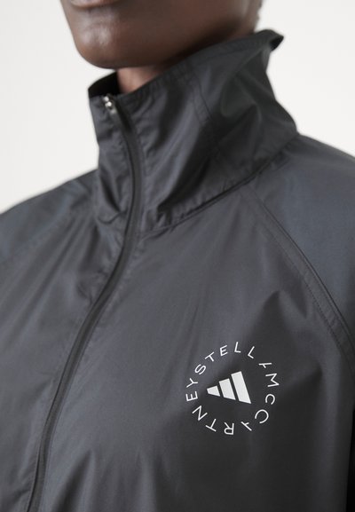 Zwarte lichte windbreaker jas met een hoge kraag, volledige rits aan de voorkant en een wit cirkelvormig logo op de linkerborst.