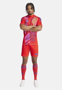 Rote Fußballausrüstung bestehend aus einem kurzärmeligen Shirt mit geometrischen Mustern in Pink und Blau. Enthält passende Shorts und Socken mit blauen Akzenten.