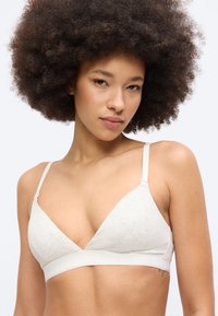 Νεαρή γυναίκα με μεγάλα φυσικά σγουρά μαλλιά που φοράει ανοιχτό γκρι, μαλακό σουτιέν με λεπτές ρυθμιζόμενες τιράντες.