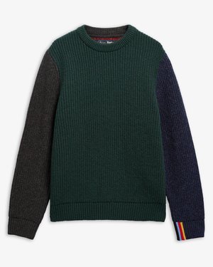 Jeune homme debout les mains dans les poches, portant un pull en maille vert foncé, une chemise à carreaux, un pantalon sombre et des chaussures marron sur fond blanc.