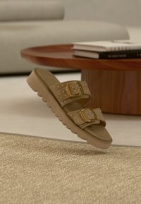 Sandale compensée marron avec deux boucles dorées et des clous dorés, placée près d'une table basse en bois avec des livres empilés dans un salon beige.