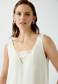 Cremfarbene ärmellose Bluse mit tiefem V-Ausschnitt, die ein Spitzenelement am Halsbereich und eine glatte Stoffstruktur aufweist.
