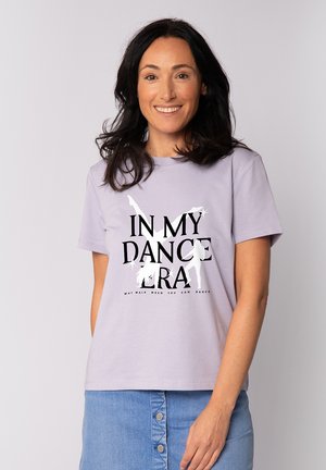 Femme aux cheveux noirs portant une chemise lilas avec l'inscription « IN MY DANCE ERA » et des silhouettes dansantes, associée à une jupe en jean bleue à boutons sur le devant.
