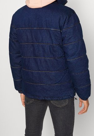 Personne portant une veste matelassée bleu foncé avec des coutures horizontales et une capuche de couleur claire, assortie à un jean gris foncé.
