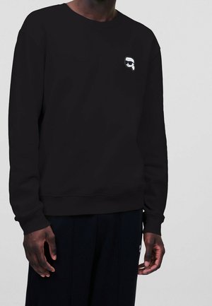 Osoba ubrana w czarną bluzę z długim rękawem, z małym biało-czarnym uśmiechniętym twarzą w okularach przeciwsłonecznych na lewej piersi.